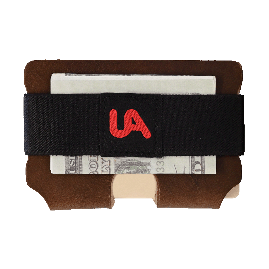 Brown leather EDC wallet with black elastic money clip featuring red UA logo, holding folded U.S. cash, on white background.