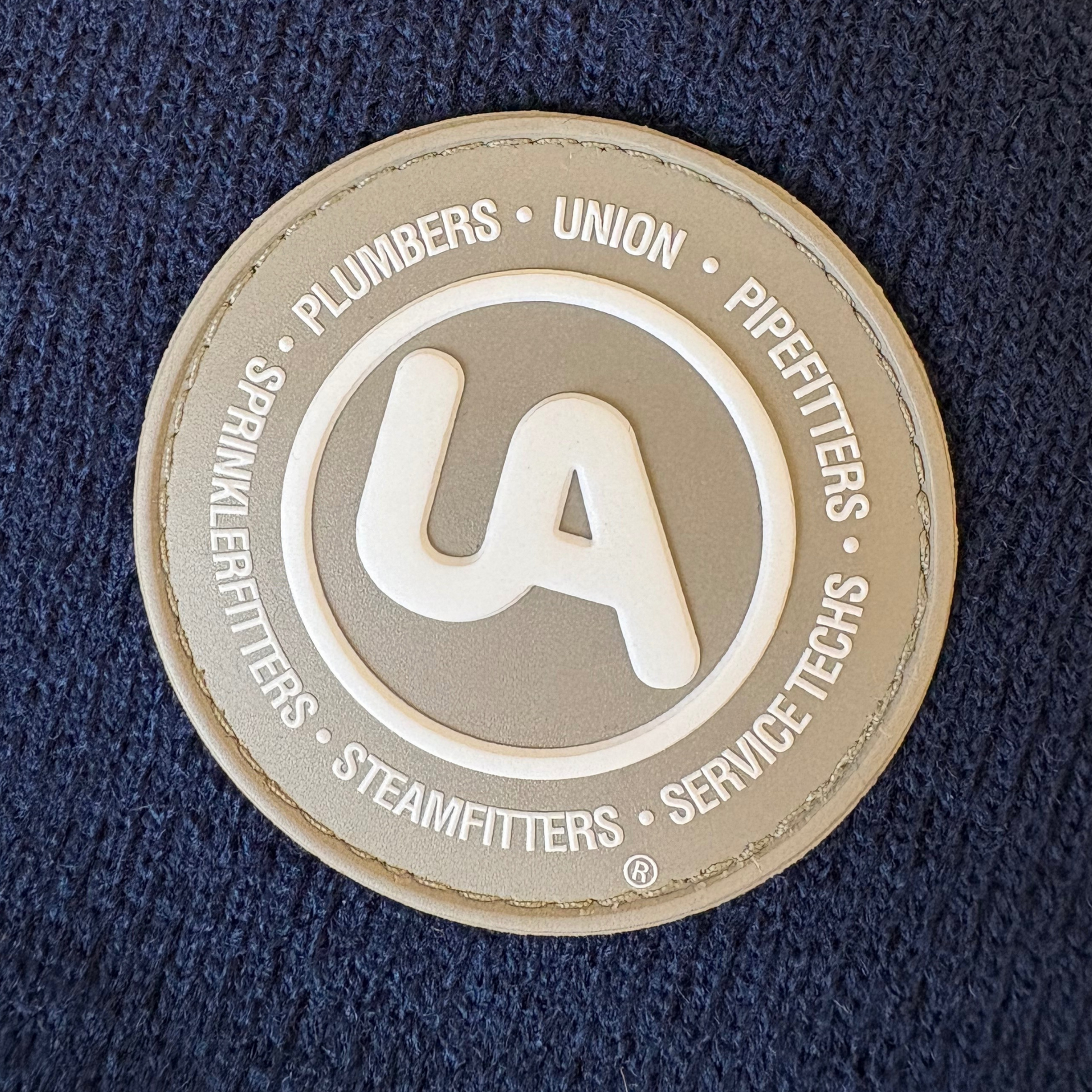 Close-up of a circular UA union patch featuring raised white text reading “Plumbers, Pipefitters, Sprinklerfitters, Steamfitters, Service Techs” on a gray background, stitched onto navy knit fabric.